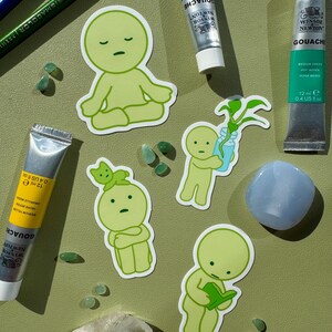 May include: Five green stickers featuring cartoon characters with different expressions. The characters are all wearing green clothing and have a simple design. The stickers are on a green background with small green and blue stones.