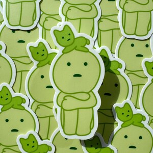 May include: A collection of green stickers featuring a cartoon character with a cat on its head. The character has a simple design with a round head, arms crossed, and legs. The cat is also simple, with a round head, ears, and a tail.