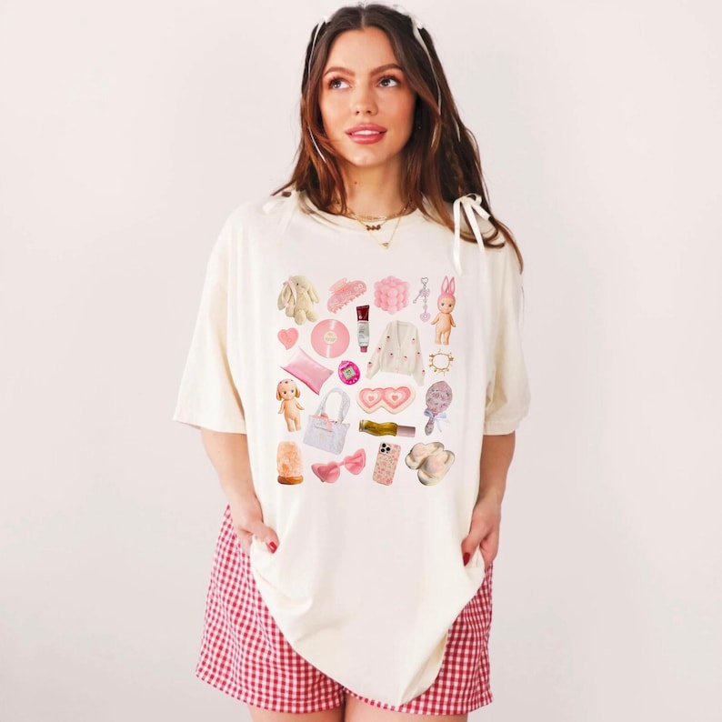 May include: A white t-shirt with a pink and white graphic print of various items, including a heart, a doll, a phone, a pair of slippers, and a pink bow.