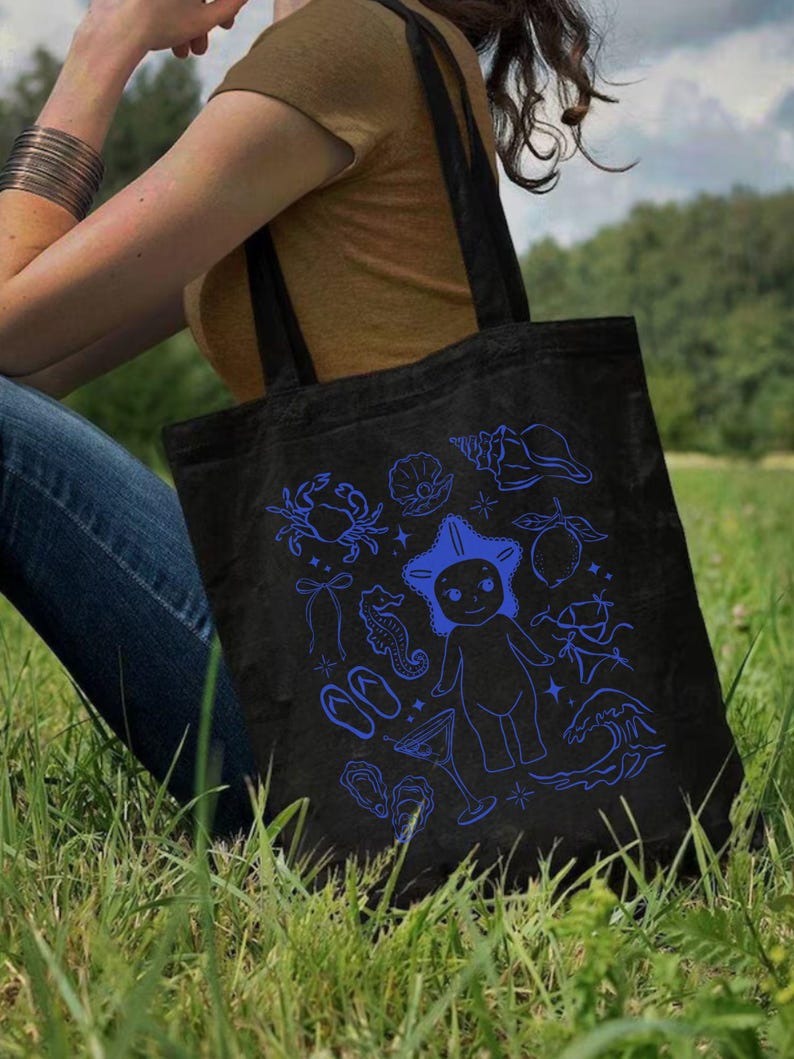 May include: Black tote bag with blue illustrations of sea creatures, including a crab, starfish, seahorse, and seashells. The bag has two black handles and is made of a sturdy fabric. The design is whimsical and nautical-themed.