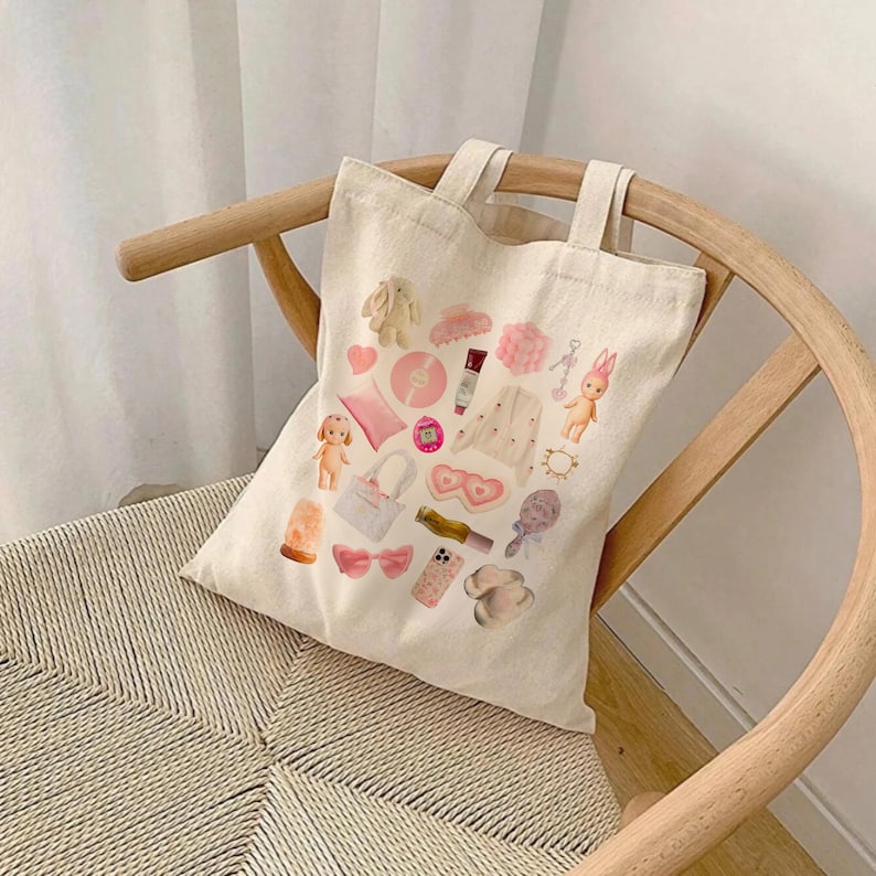 May include: A white canvas tote bag with a pink and white design featuring various items, including a pink heart, a pink bunny, a pink phone, a pink hair clip, a pink compact, a pink lipstick, a pink ring, a pink flower, a pink doll, a pink lamp, a pink bow, a pink bag, a pink eye mask, a pink headband, and a pink bracelet.