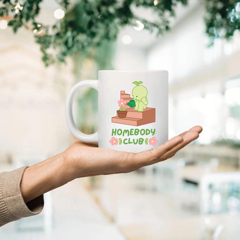 May include: White ceramic mug featuring a cartoon illustration of a green character watering a plant on a brown structure. The mug has the text "HOMEBODY CLUB" in green and pink. The mug is held in a hand.