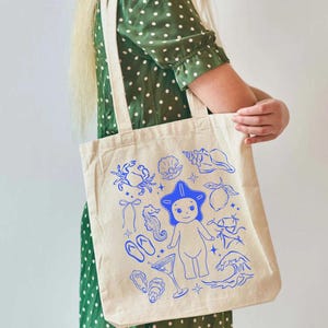 May include: A beige tote bag with blue illustrations of beach-themed elements, including a crab, seashells, a seahorse, a martini glass, and a cartoon figure with a star-shaped hat. The bag has two cream-colored handles.