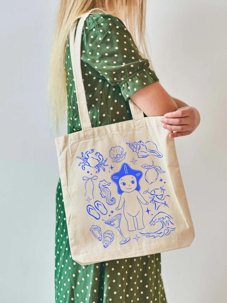 May include: A beige tote bag with blue illustrations of beach-themed elements, including a crab, seashells, a seahorse, a martini glass, and a cartoon figure with a star-shaped hat. The bag has two cream-colored handles.
