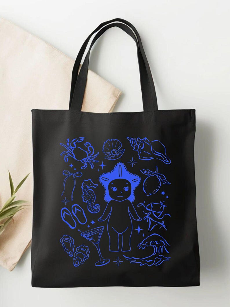 May include: Black tote bag with a blue illustration of beach-themed elements. The design includes a crab, seashells, a starfish-headed figure, a seahorse, flip-flops, a cocktail, and a wave.
