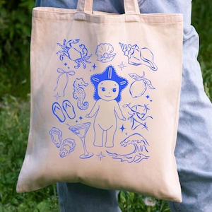 May include: Beige tote bag with a blue illustration of a starfish-headed figure, seashells, a crab, and other beach-themed elements. The bag has two handles and is made of a canvas-like material.