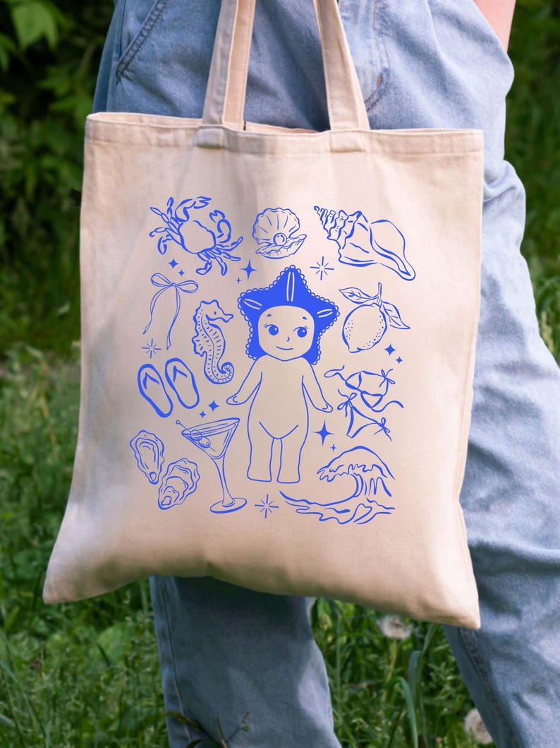 May include: Beige tote bag with a blue illustration of a starfish-headed figure, seashells, a crab, and other beach-themed elements. The bag has two handles and is made of a canvas-like material.