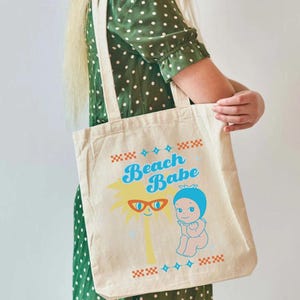 May include: A beige canvas tote bag with the words "Beach Babe" in blue and yellow. The bag features a cartoon palm tree wearing sunglasses and a baby in a blue bonnet. The bag is held by a person wearing a green dress.
