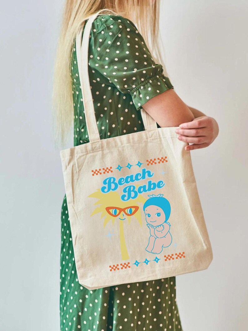May include: A beige canvas tote bag with the words "Beach Babe" in blue and yellow. The bag features a cartoon palm tree wearing sunglasses and a baby in a blue bonnet. The bag is held by a person wearing a green dress.