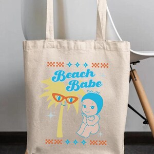May include: Beige tote bag with the words "Beach Babe" in blue script. The bag features a cartoon palm tree wearing sunglasses and a baby in a blue bonnet. The design includes orange and blue decorative elements, perfect for carrying beach essentials.