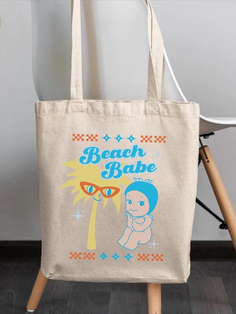 May include: Beige tote bag with the words "Beach Babe" in blue script. The bag features a cartoon palm tree wearing sunglasses and a baby in a blue bonnet. The design includes orange and blue decorative elements, perfect for carrying beach essentials.