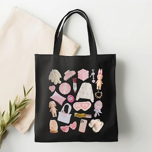 May include: Black canvas tote bag with a pink and white design featuring various items like a bunny, a hair clip, a heart, a pillow, a doll, a headband, a phone case, a pair of slippers, and a makeup product.