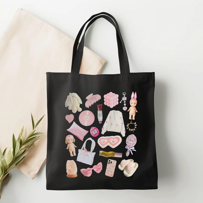 May include: Black canvas tote bag with a pink and white design featuring various items like a bunny, a hair clip, a heart, a pillow, a doll, a headband, a phone case, a pair of slippers, and a makeup product.