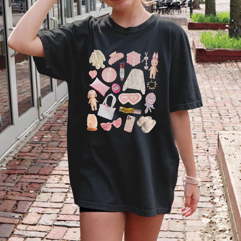 May include: A black t-shirt with a pink and white graphic print of various items, including a doll, a purse, a phone, a bow, and a pair of eye masks.