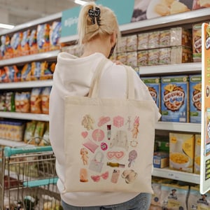 May include: A white tote bag with a variety of colorful, cartoon-like images and designs. The bag is decorated with images of hearts, flowers, a bunny, a doll, and other whimsical elements. The bag is being held by a person in a grocery store.