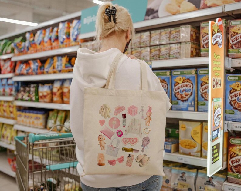 May include: A white tote bag with a variety of colorful, cartoon-like images and designs. The bag is decorated with images of hearts, flowers, a bunny, a doll, and other whimsical elements. The bag is being held by a person in a grocery store.