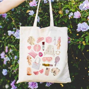 May include: A white tote bag with a pink and white design featuring various items, including a bunny, a heart-shaped eye mask, a phone, a hair clip, a doll, and a salt lamp.
