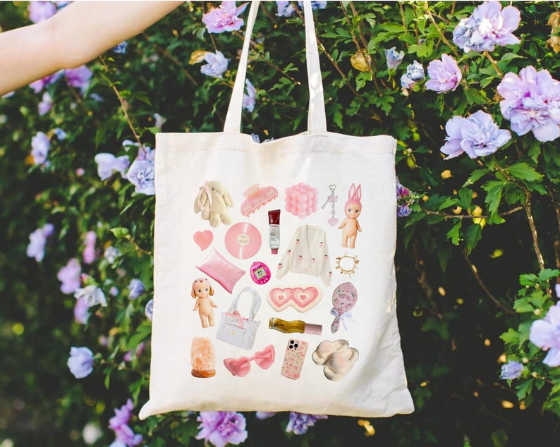 May include: A white tote bag with a pink and white design featuring various items, including a bunny, a heart-shaped eye mask, a phone, a hair clip, a doll, and a salt lamp.