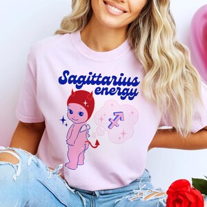 Puede incluir: Camiseta rosa con un diablo de dibujos animados con cuernos rojos y un s&iacute;mbolo de Sagitario azul y rojo en una nube rosa. El texto "Sagittarius energy" est&aacute; escrito en letras azules y rojas.
