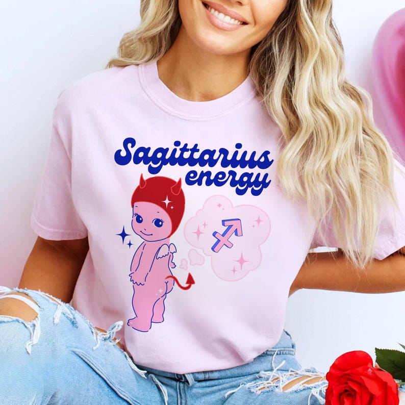 Puede incluir: Camiseta rosa con un diablo de dibujos animados con cuernos rojos y un s&iacute;mbolo de Sagitario azul y rojo en una nube rosa. El texto "Sagittarius energy" est&aacute; escrito en letras azules y rojas.
