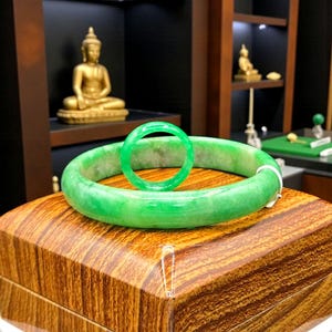 Könnte beinhalten: Ein leuchtend grünes Jadeit-Jade-Armreif- und Ring-Set. Das Jade hat eine lebendige grüne Farbe mit einem leicht durchscheinenden Aussehen. Das Set wird auf einer Holzkiste mit detailliertem Maserungsmuster präsentiert.