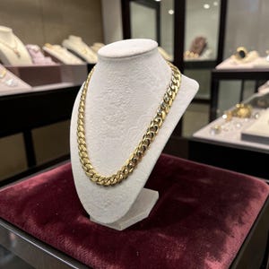 May include: A thick, polished gold-tone curb chain necklace featuring interlocking, rounded metal links. The necklace is displayed on a neutral-colored, fabric-covered bust stand, showcasing the uniform size and reflective surface of each individual link.
