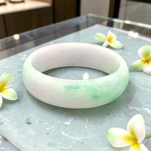 May include: A jade bangle bracelet with a smooth, polished surface. The bracelet features a gradient of colors, transitioning from white to a soft green. The bracelet is displayed on a light gray surface, adorned with white and yellow flowers.