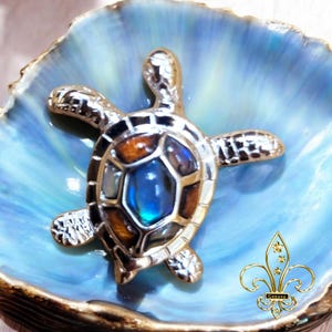 May include: A silver turtle pendant with a blue and brown iridescent shell. The turtle is sitting on a blue and white seashell.