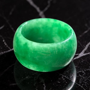 Puede incluir: Un anillo de banda ancha y sólida hecho de jade verde pulido. El anillo presenta una superficie exterior lisa y redondeada con variaciones naturales de color, que van desde el verde esmeralda profundo hasta tonos más claros y translúcidos.