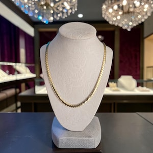 Puede incluir: Un collar de cadena de oro con un diseño de eslabones planos entrelazados en forma de espiga. El collar se exhibe sobre un busto de color neutro y textura, mostrando el acabado metálico pulido y el patrón repetitivo de los eslabones.