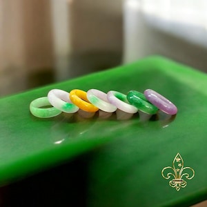 May include: A set of six jade rings in various shades of green, yellow, and purple. The rings are arranged in a row on a green surface.