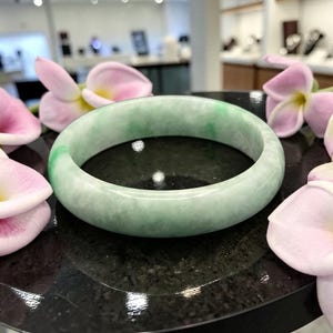 May include: A round jade bangle bracelet with a light green hue and subtle variations in color. The polished surface reflects light, and the bracelet is displayed on a dark surface with pink flowers.