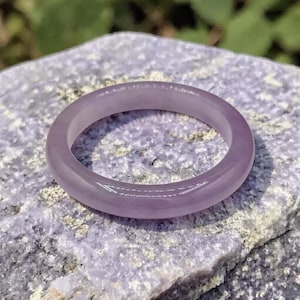 May include: A translucent, lavender-colored ring. The ring is made of a smooth, polished material and is set against a textured, light purple stone surface. The ring's color is consistent throughout, with a subtle sheen.