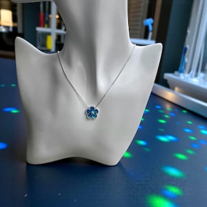May include: A silver necklace with a blue and turquoise flower pendant. The flower has a small, clear gem in the center. The necklace is displayed on a white mannequin bust against a blue background.