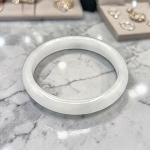 Puede incluir: Un brazalete de jade blanco. El brazalete liso y redondo es de color blanco opaco sólido. La joya se exhibe sobre una superficie de mármol, con otras piezas de joyería visibles en el fondo.