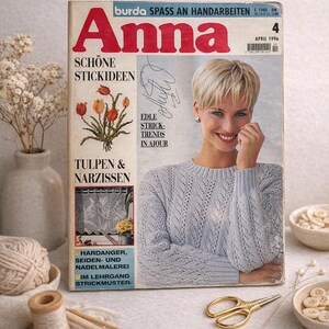 May include: A vintage magazine titled "Anna" with a woman on the cover wearing a light blue sweater. The magazine features articles on knitting and embroidery. A vase of flowers, yarn, needles, and scissors are also visible.