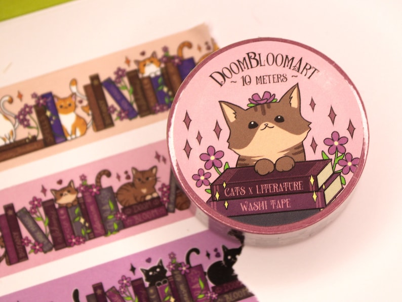 May include: A pink and purple washi tape roll featuring a cat sitting on a stack of books. The tape is labelled "Doombloomart - 10 metres - Cats x Literature Washi Tape".