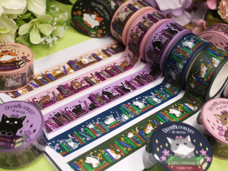 May include: A selection of colourful washi tapes featuring cats reading books. The tapes have a variety of designs, including purple, blue, green and pink. The text on the tapes reads "Doombloomart" and "10 metres".