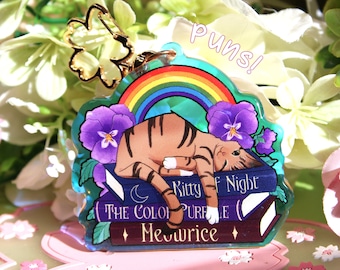Op de afbeelding: Een regenboog- en paarse bloemenbedel met een slapende kat op een stapel boeken. De boeken zijn getiteld "Kitty F Night", "The Color Purple" en "Meowrice".