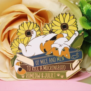 Puede incluir: Un pin de esmalte dorado con un gato acostado sobre una pila de libros. Los libros tienen los títulos "Of Mice and Men", "To Kill a Mockingbird" y "Romeow & Juliet". El gato es blanco con marcas naranjas y negras. El gato está rodeado de girasoles amarillos.