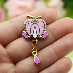 Könnte beinhalten: Goldfarbene Emaille-Anstecknadel mit einer rosa Blume mit weißem Zentrum und einem herabhängenden violetten Tropfen. Die Blume hat einen goldfarbenen Stiel und Blätter.