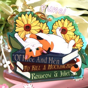Op de afbeelding: Een heldere acryl sleutelhanger met een lapjeskat die op een stapel boeken ligt met zonnebloemen. De boeken heten "Of Mice And Men", "To Kill A Mockingbird" en "Romeow & Juliet". Het woord "Puns!" staat bovenaan.