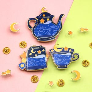 May include: Enamel pins with a celestial tea party design. The set includes a blue teapot, teacup, and teabag, decorated with moons, stars, and clouds. Additional pins include gold-coloured stars, moons, and round pin backs.