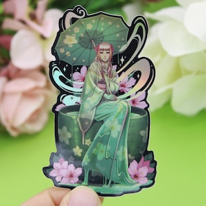 Könnte beinhalten: Ein Aufkleber mit einer Frau in einem grünen Kimono, die auf einer grünen Teetasse sitzt. Sie hält einen Regenschirm mit rosa Blumen und ist von rosa Blumen umgeben. Der Aufkleber hat eine holografische Oberfläche.