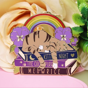 Könnte beinhalten: Goldene Emaille-Anstecknadel mit einer braunen Tabby-Katze, die auf einem Stapel Bücher schläft. Die Bücher tragen die Titel "The Color Purple", "Kitty Night" und "Meowrice". Ein Regenbogen befindet sich über der Katze und violette Stiefmütterchen befinden sich an den Seiten.