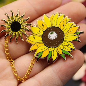 Op de afbeelding: Een gouden ketting verbindt twee emaille zonnebloemspelden. De ene zonnebloem heeft een zwart midden, terwijl de andere een bruin midden heeft met een wit oog.