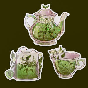 May include: Set of three stickers featuring tea-themed illustrations. Includes a teapot, a teabag, and a teacup, all filled with green liquid and floral accents. The stickers have a white border and a glittery effect.