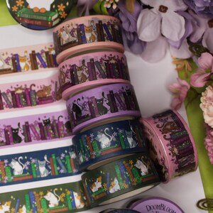 May include: A stack of six colourful washi tapes featuring cats and books. The tapes are decorated with purple, pink, blue, and green colours. The cats are depicted in various poses, including sitting, standing, and sleeping. The books are stacked on top of each other and have different colours and designs.