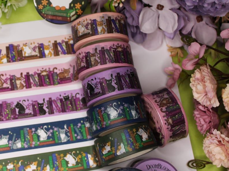 May include: A stack of six colourful washi tapes featuring cats and books. The tapes are decorated with purple, pink, blue, and green colours. The cats are depicted in various poses, including sitting, standing, and sleeping. The books are stacked on top of each other and have different colours and designs.