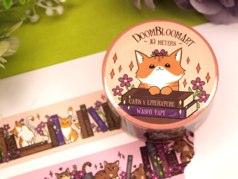 May include: A roll of pink and orange washi tape featuring a cute orange cat illustration. The tape has a stack of books with the text "Cats x Literature" and "Washi Tape". The tape is 10 metres long.